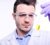 Mann im Laborkittel mit Handschuhen und Schutzbrille schaut auf Reagenzglas mit gelber Flüssigkeit